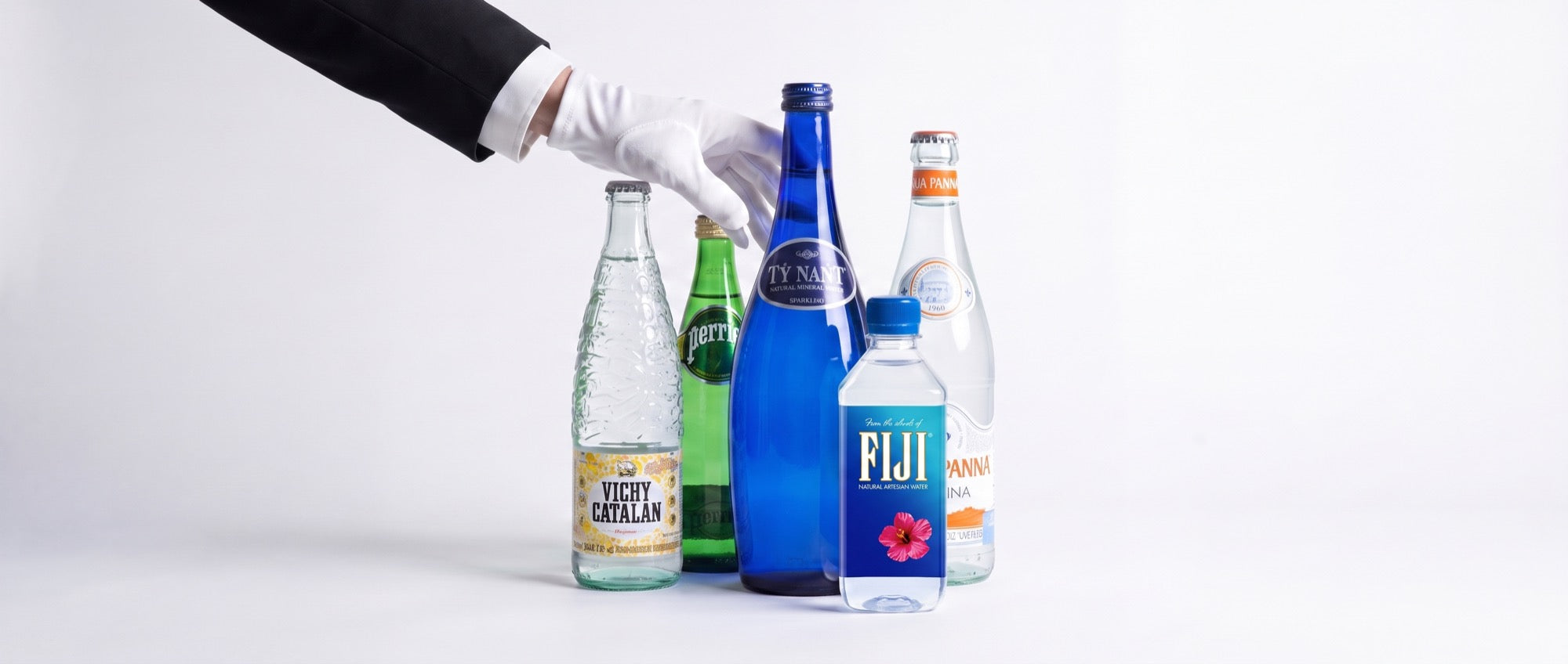 A white-gloved hand selects from five premium bottled waters — Vichy Catalan, Perrier, Tŷ Nant, Fiji and Acqua Panna — arranged on a pale table